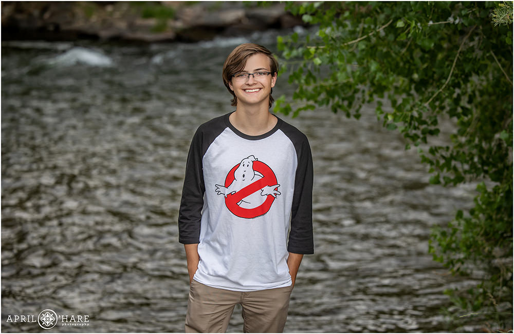 Young adult man wearing a baseball style ghostbusters tshirt stands in front of Clear Creek near the Clear Creek History Park in Golden Colorado