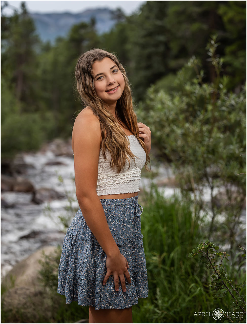 High school senior girl with long wavy hair poses with the Big Thompson River in the backdrop at Rocky Mountain National Park