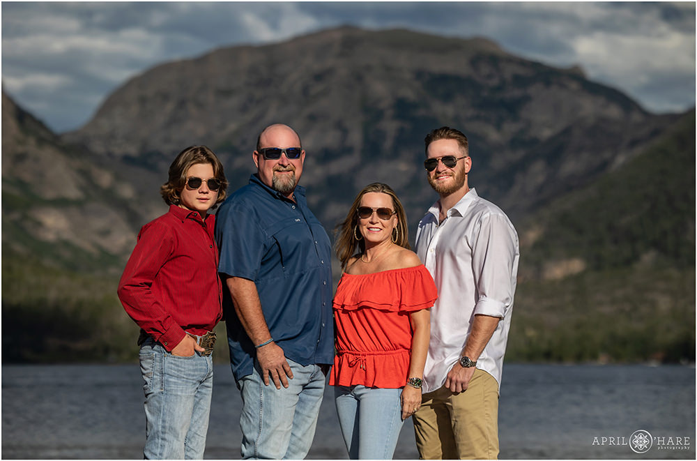 Family of four with two older sons all wear sunglasses in front of the pretty mountain backdrop at Grand Lake in Colorado