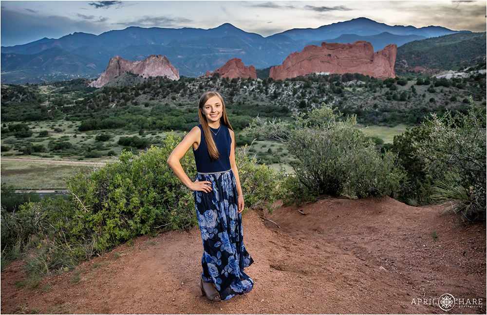 Teen girl wearing a long blue floral dress poses at Mesa Overlook with Garden of the Gods in the backdrop