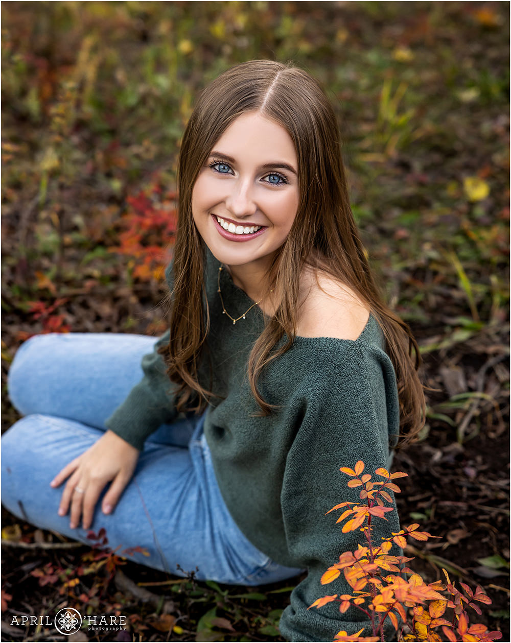 Senior girl with bright blue eyes wearing a green sweater and jeans with some pretty fall color framing her