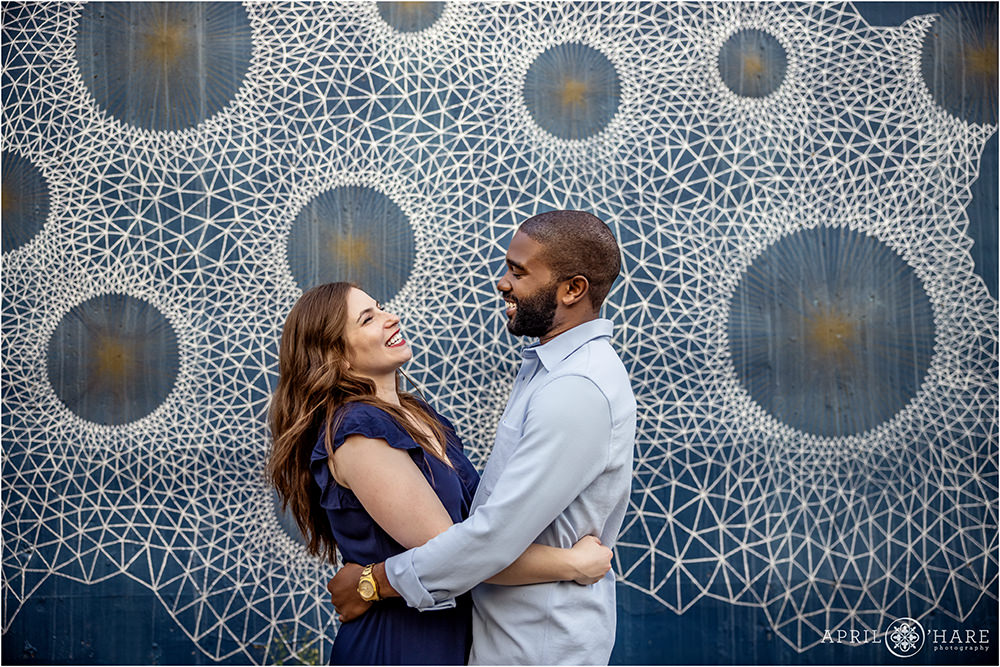 Cute couple laugh together in front of a pretty blue geometric backdrop along Cherry Creek in Denver