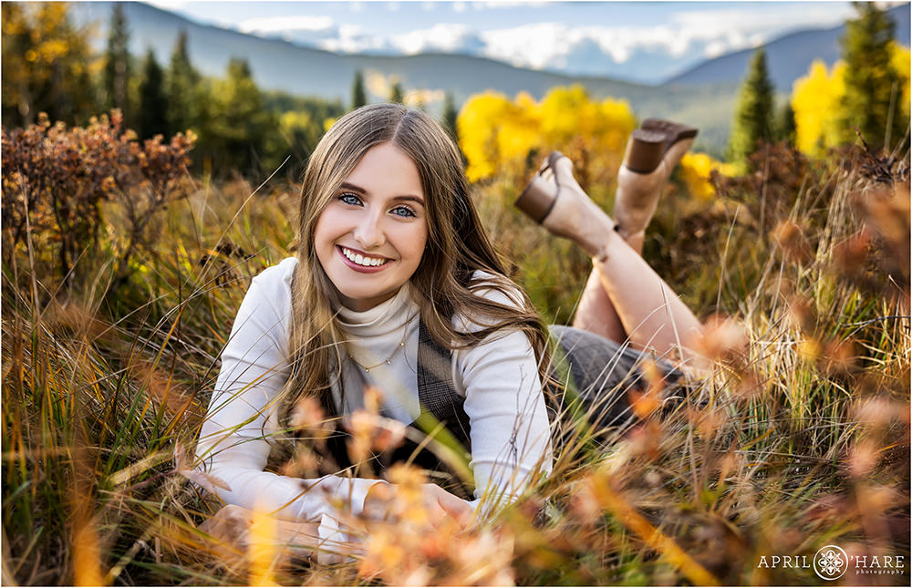 High school senior girl lies in a mountain meadow with pretty mountain backdrop and aspen trees in the distance