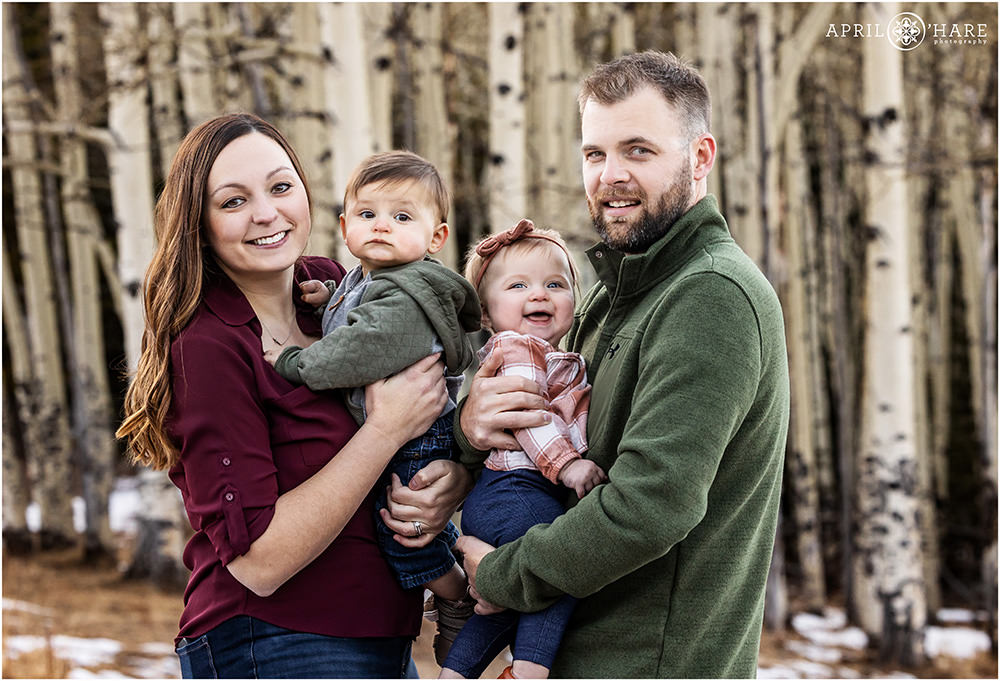 Sweet baby twins are held by their parents with aspen tree backdrop during winter in Colorado