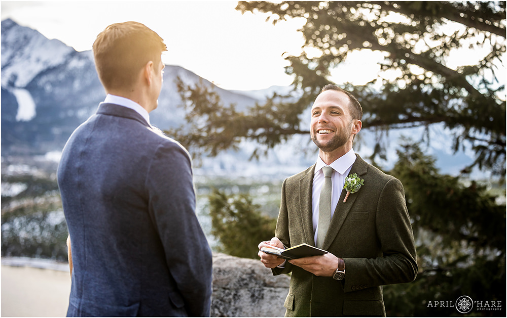 Groom wearing olive green suit smiles at his groom as he says his vows at Sapphire Point