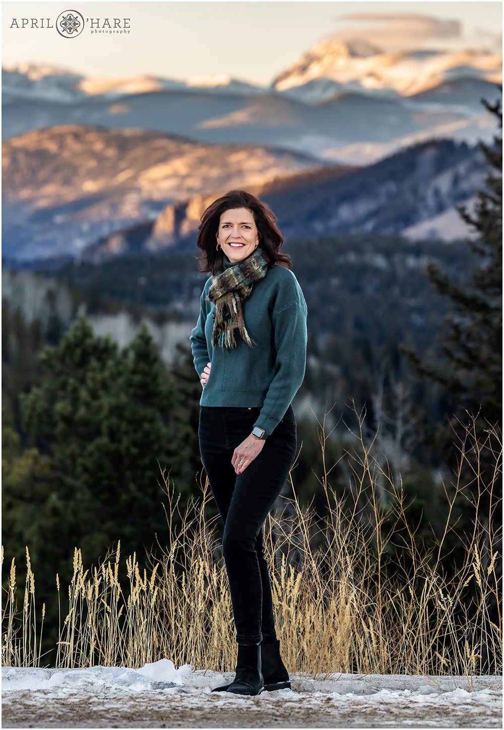 A woman wearing a teal sweater with a scarf poses with a beautiful mountain backdrop behind her during winter in Evergreen Colorado