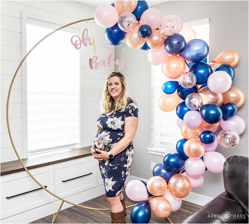Cute photo of a pregnant woman wearing a blue floral dress standing in front of her blue and pink balloon arch backdrop at her baby shower in Colorado
