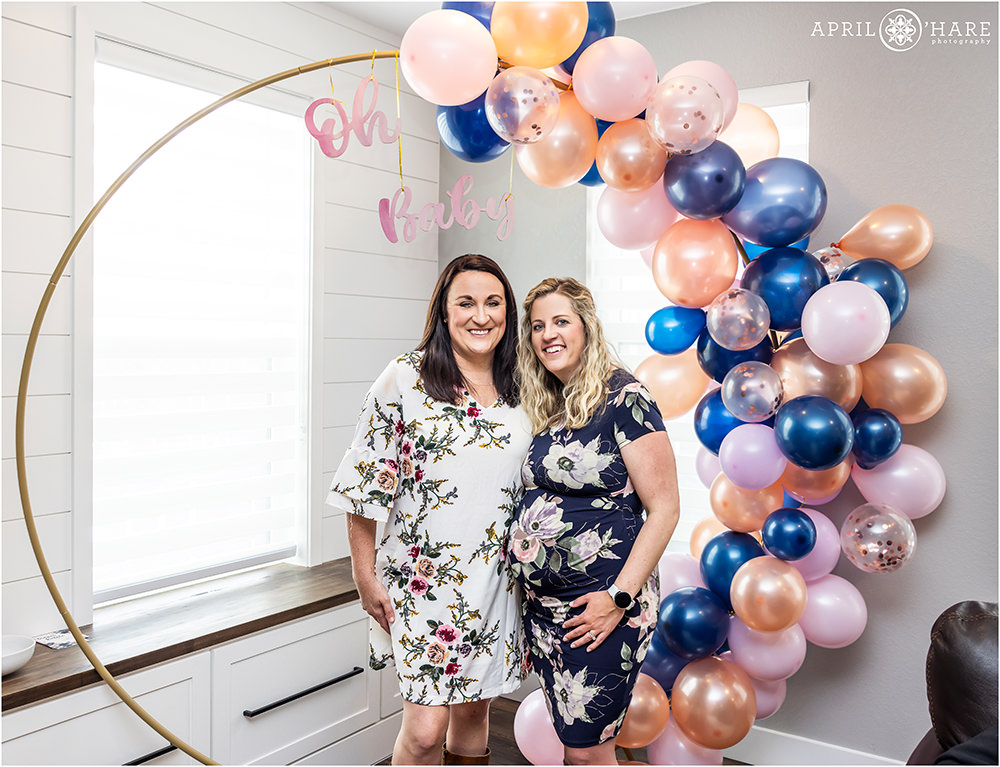 Cute lesbian couple pose in front of a blue and pink balloon arch at their baby shower in Littleton Colorado