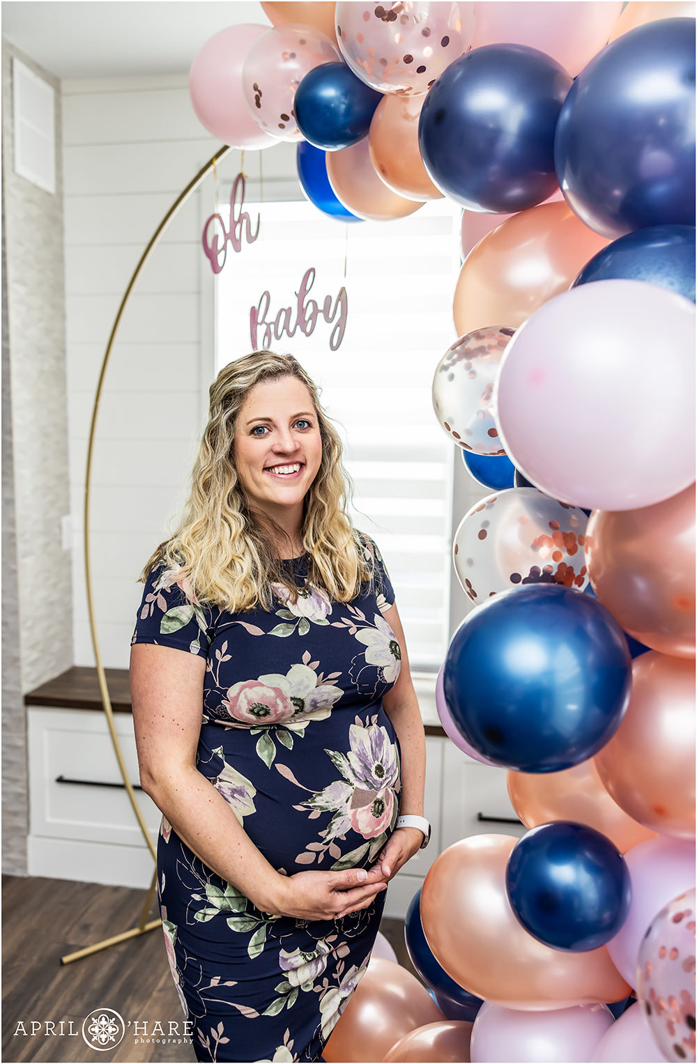 Colorado baby shower photographer photographs a mama to be wearing a blue and pink floral dress at her blue and pink themed baby shower as she stands in front of her cute blue and pink balloon arch