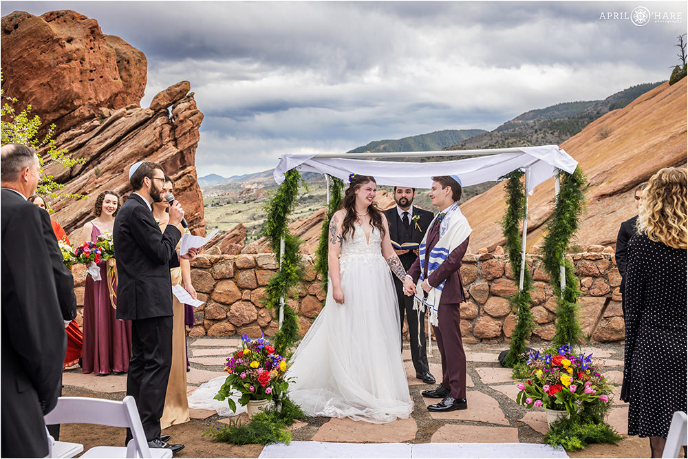 Bride and groom smile at each other as their best man gives a reading during their wedding ceremony at Red Rocks