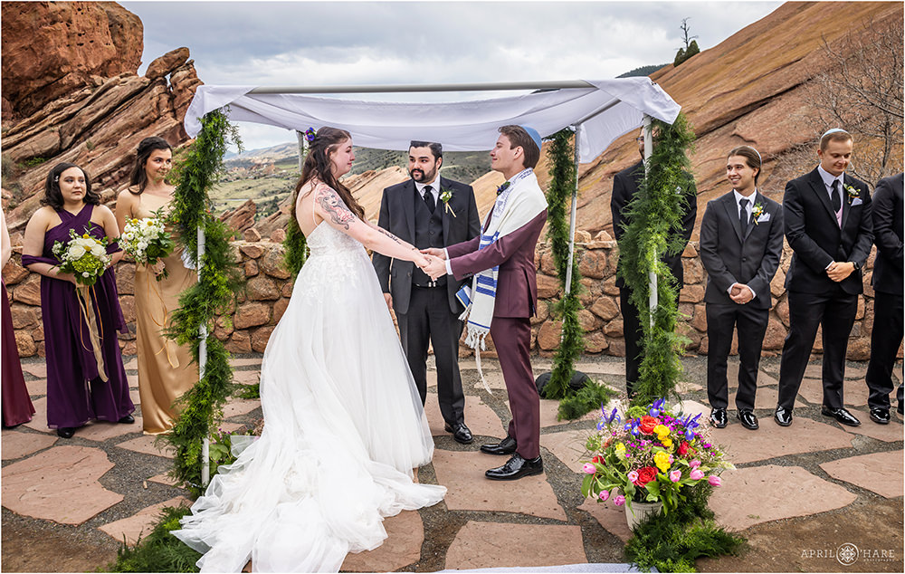 Bride and groom hold hands and look at each other as they get married under the huppah at Red Rocks