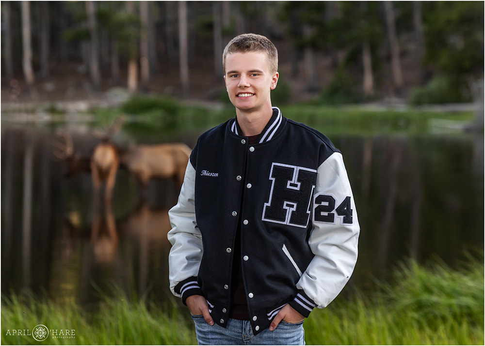 High school senior portrait of a young man wearing a letter jacket with two elk sipping water at Lake Sprague behind him