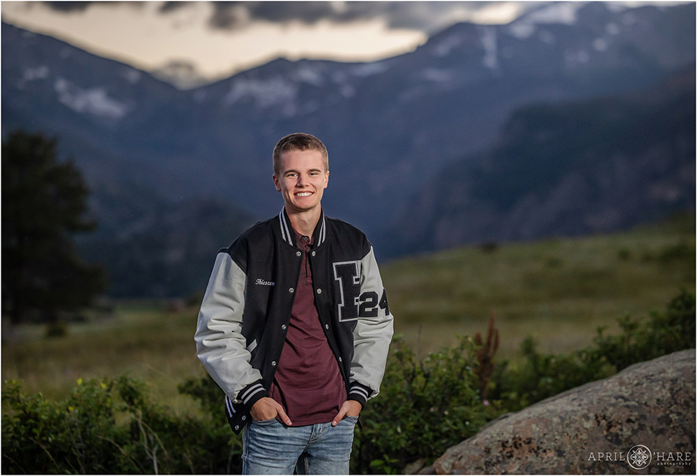A young man poses for a high school senior photo in his letter jacket at Moraine Park with the mountain view behind him at sunset in Colorado