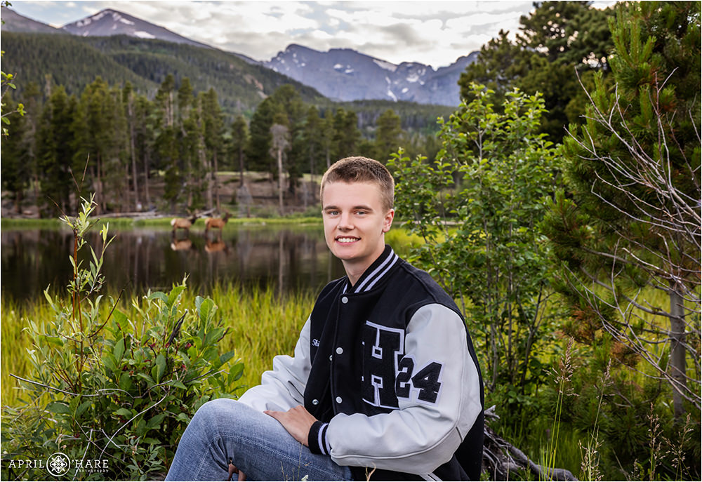 Senior photo with a young man wearing a high school letter jacket with two elk posing behind him in the distance