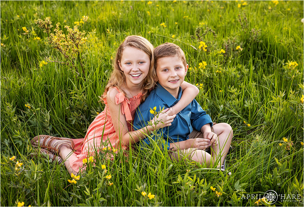 Sister with her brother sit in a grassy mountain meadow with pretty yellow wildflowers in Colorado in summer