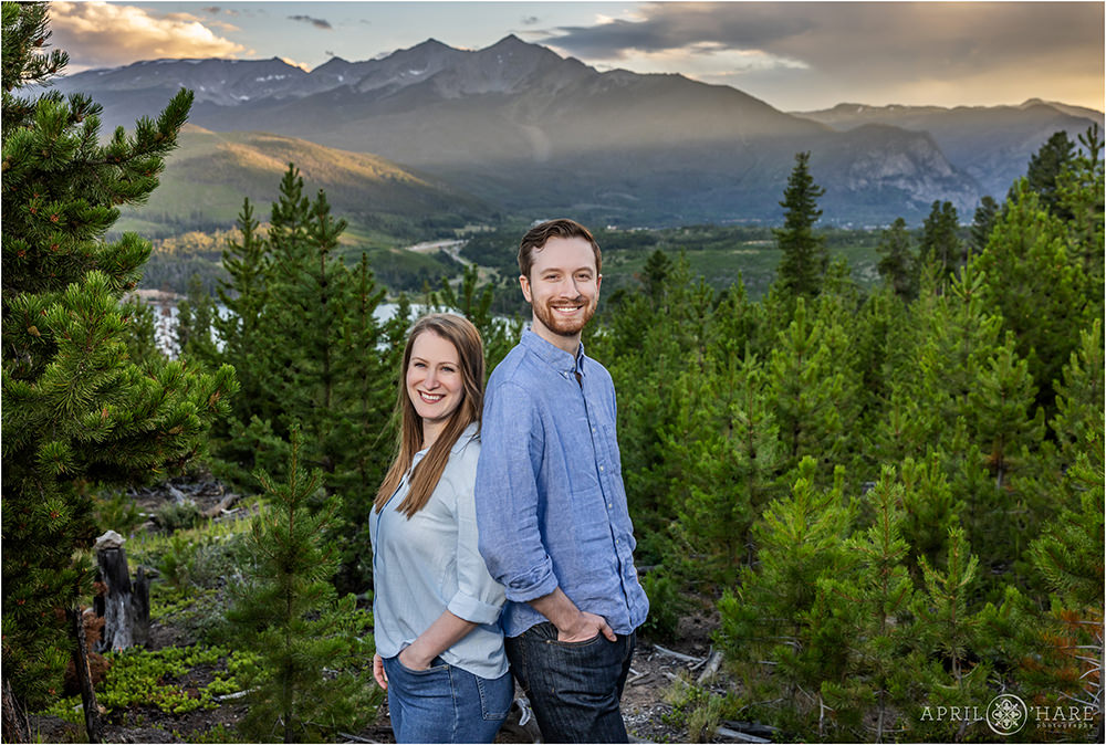Adult twin siblings stand back to back in front of a pretty mountain backdrop at Sunset in Colorado