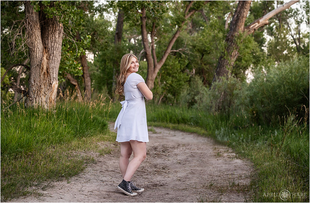 Senior girl wearing a short white dress and black converse sneakers walks along a tree lined path in Parker Colorado