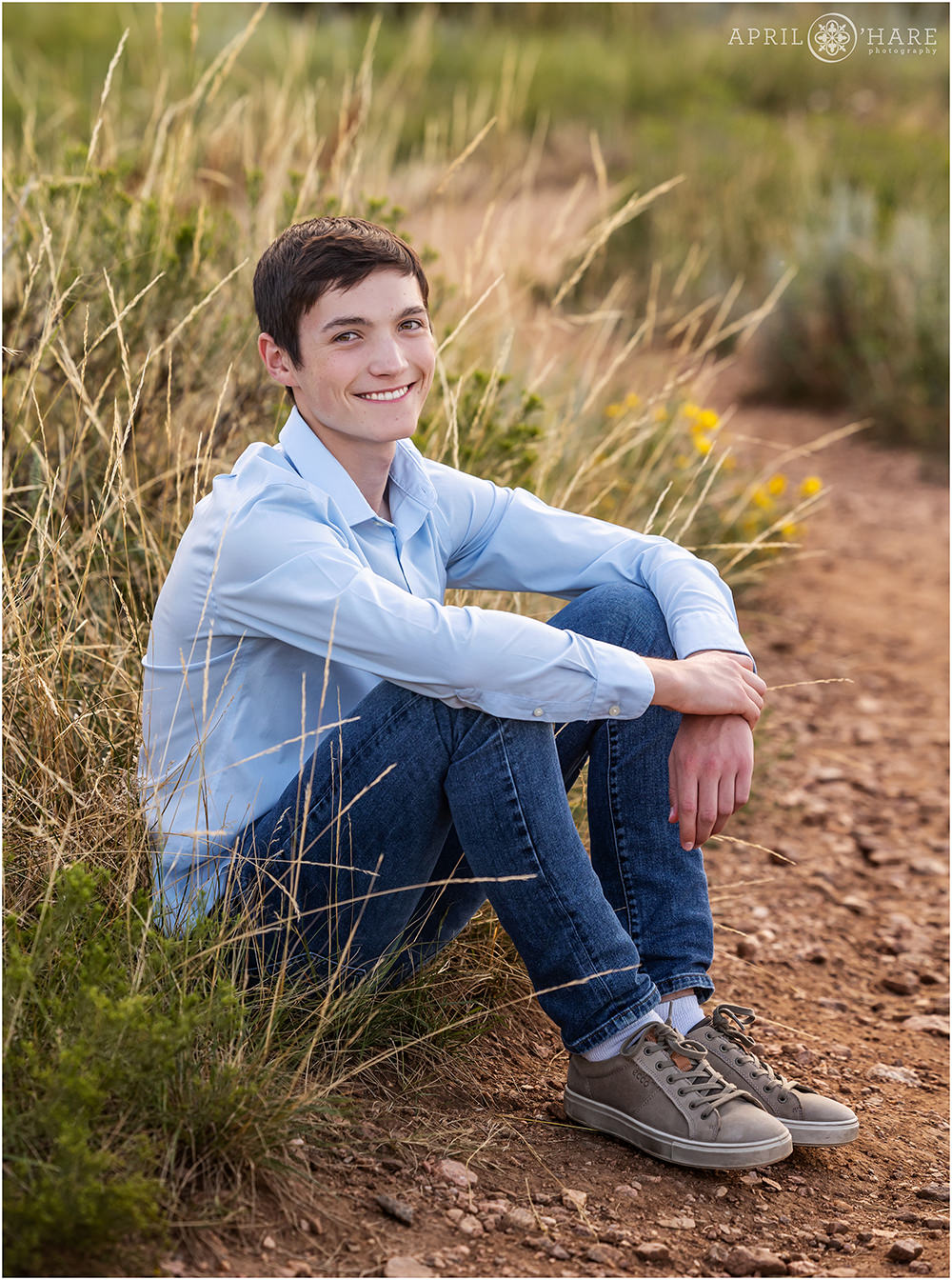 Young senior guy sits on the edge of the trail with pretty tall grasses and yellow wildflowers in the backdrop in Morrison Colorado