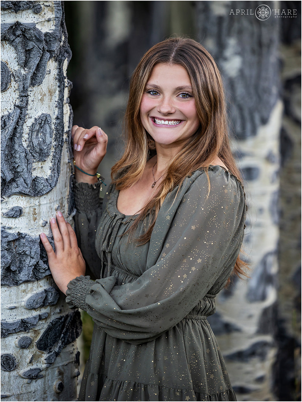 Senior Girl wearing a pretty sparkly green dress with gold on it stands next to a strand of aspen tree trunks at her Colorado high school senior photoshoot