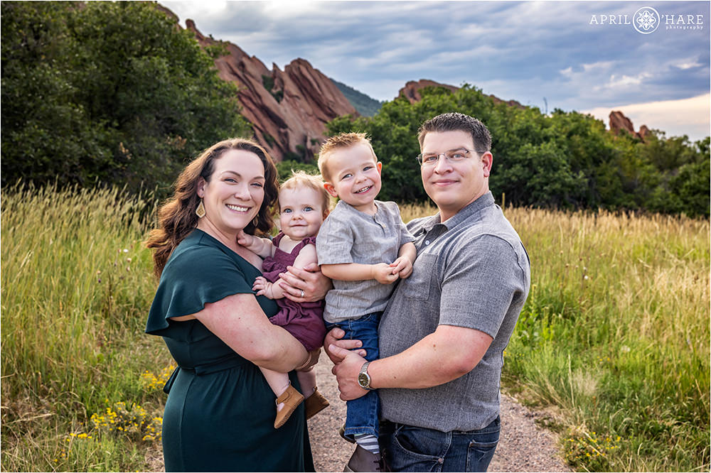 A cute family of four with a young baby daughter and a toddler son stand on the trail at Roxborough State Park in Colorado