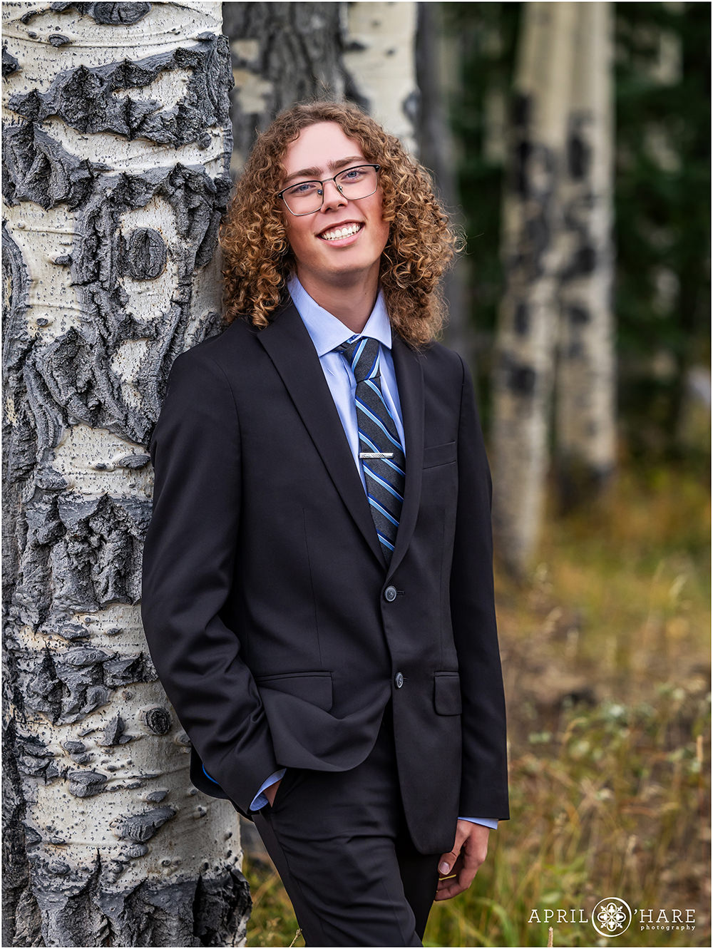Senior boy with curly hair wearing glasses and a black suit leans against an aspen tree in Evergreen Colorado