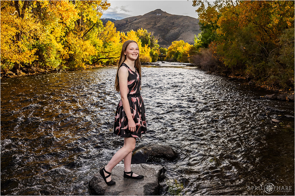 Teen girl wearing a cute black dress with a bird print on it stands in front of the gorgeous fall color Clear Creek backdrop at her senior session in Golden Colorado