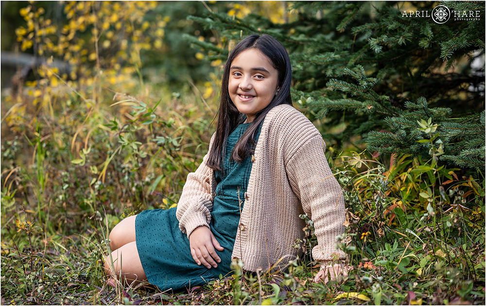 Girl wearing a green dress and tan cardigan sits in the woods with fall color in the backdrop at Squaw Pass Road in Evergreen CO