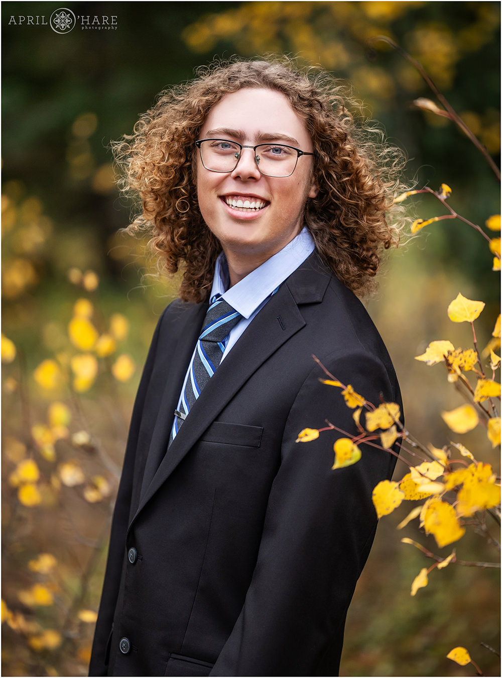 Senior boy with curly hair wearing a black suit stands in the fall color in Evergreen CO