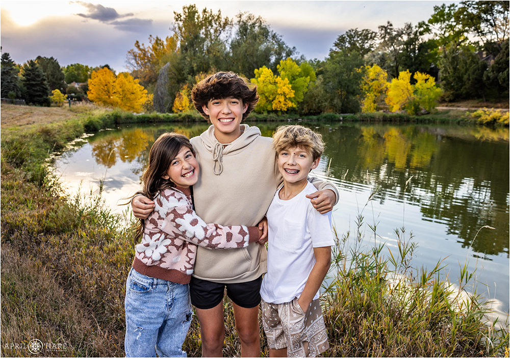 Three siblings stand in front of the pretty pond with fall color in the backdrop near their home in Littleton CO