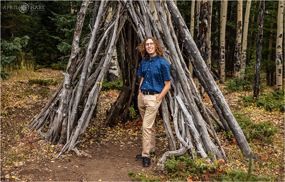 Senior boy wearing khakis with a nice blue button down shirt stands in front of a wooden tipi structure during fall in Evergreen Co