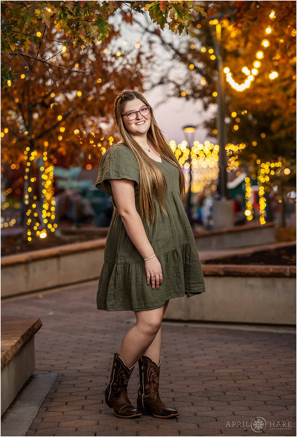 Fort Collins Senior Photographer photographs a senior girl wearing a cute green dress with cowgirl boots with the string light and fall color backdrop in Old Town Fort Collins, Colorado