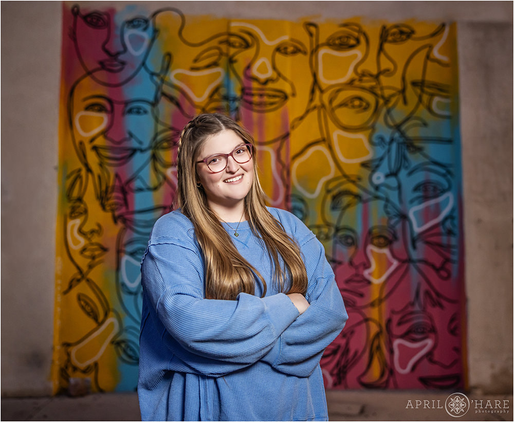 Street art wall mural with faces provides a colorful backdrop for a high school senior girl wearing a pretty blue sweater at her Fort Collins Senior photoshoot in Colorado