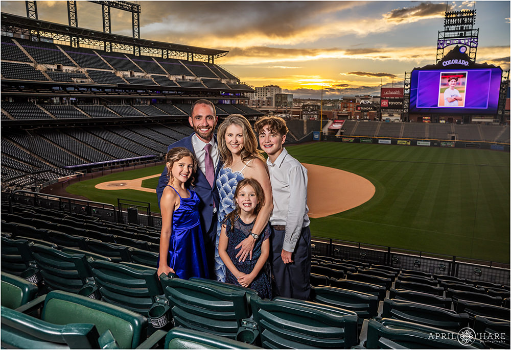 A family pose together at sunset with the Rockies scoreboard lit up and the baseball diamond backdrop at their son's bar mitzvah party at Coors Field in Denver