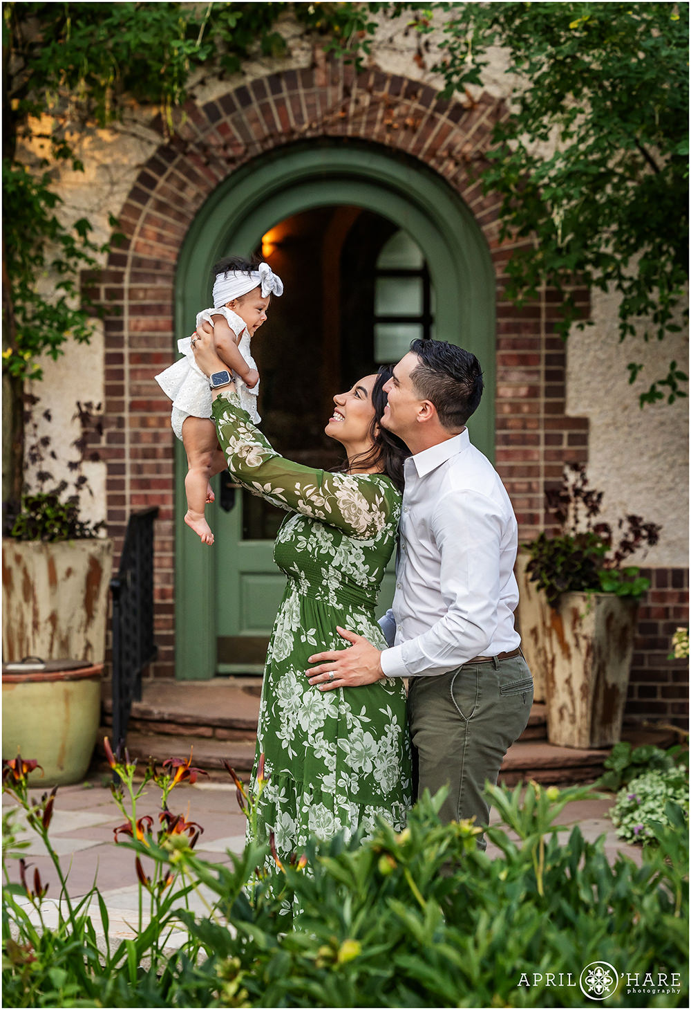 Adorable candid photo of a family of three as they lift their baby girl up in the air with a pretty arched doorway backdrop behind them at Denver Botanic Gardens