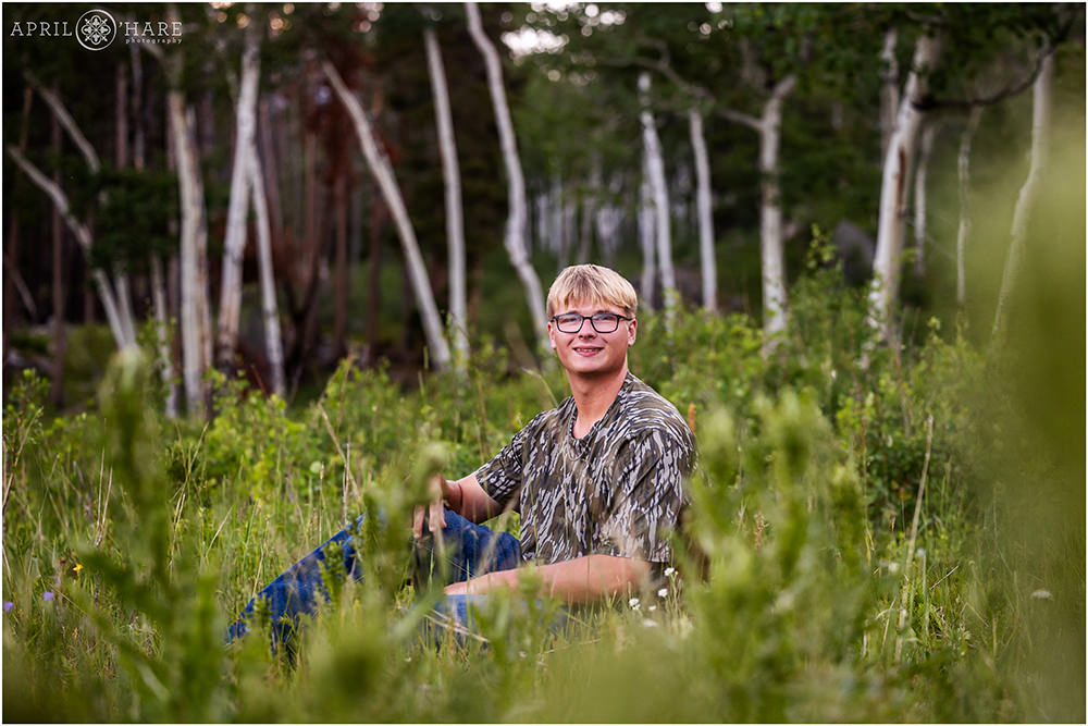 Senior portrait of a young man wearing camo in a field with grasses and aspen tree backdrop at Rocky Mountain National Park
