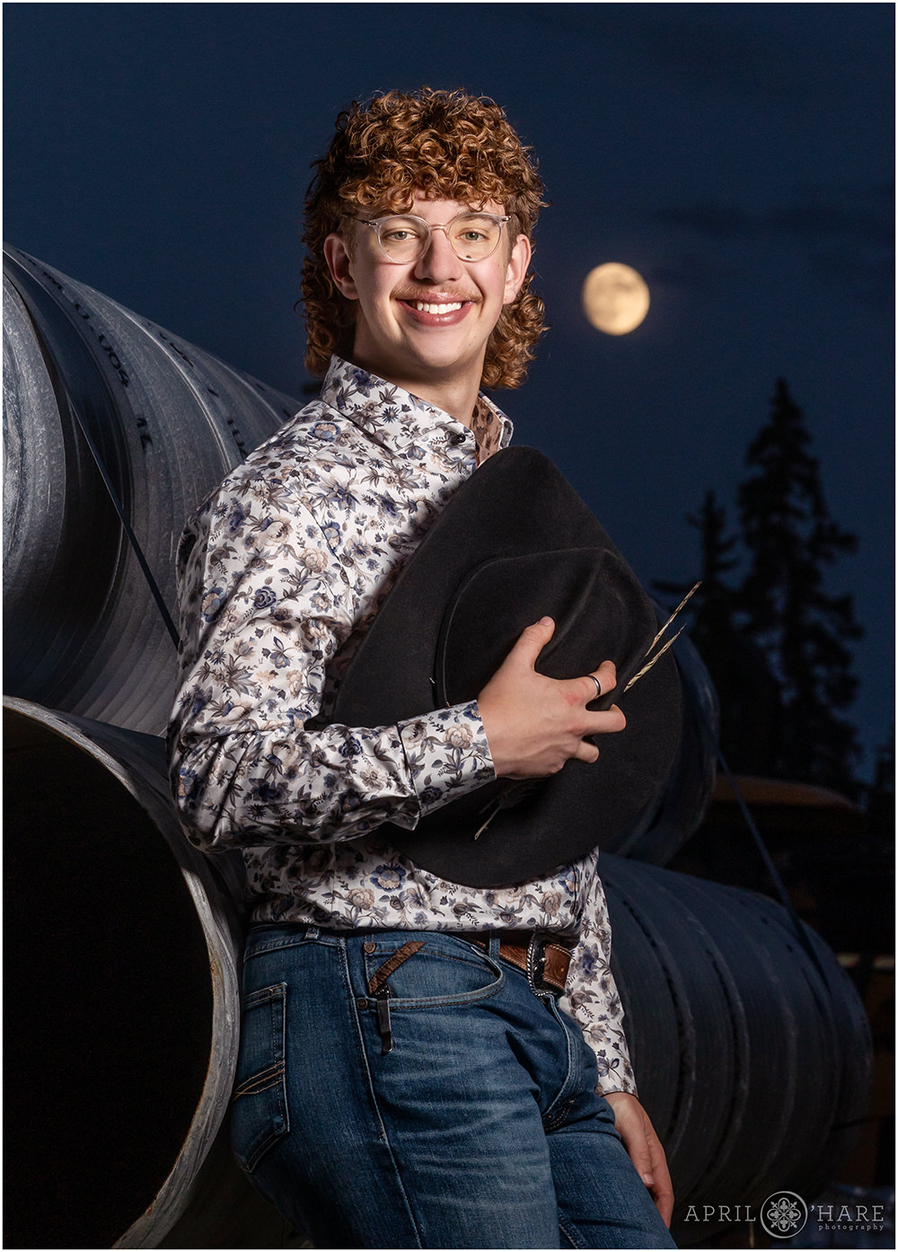 High school senior boy wearing western wear and holding his cowboy hat gets a portrait with a glowing moon behind him in the sky.