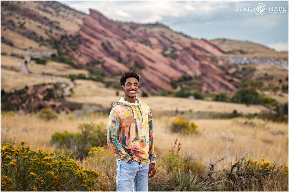 Senior boy wearing a cool artistic pain splatter style hoodie in the yellow fall colors at Morrison Trailhead with Red Rocks Amphitheater in the backdrop