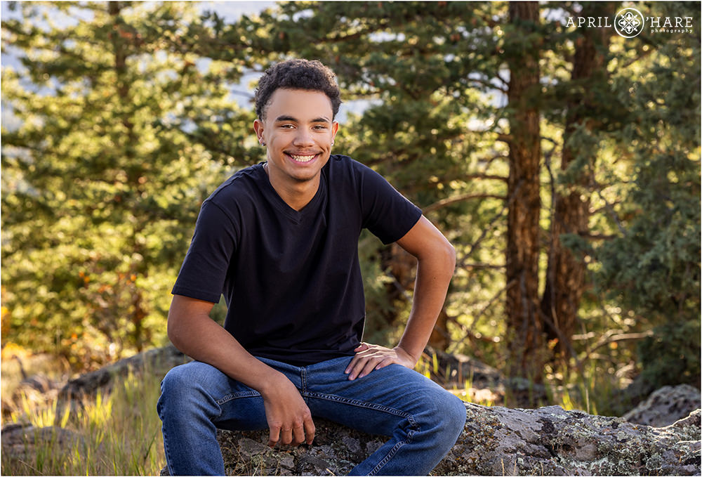 High school senior boy wearing jeans and a black tee sits on a rock for his senior portrait at West Mount Falcon