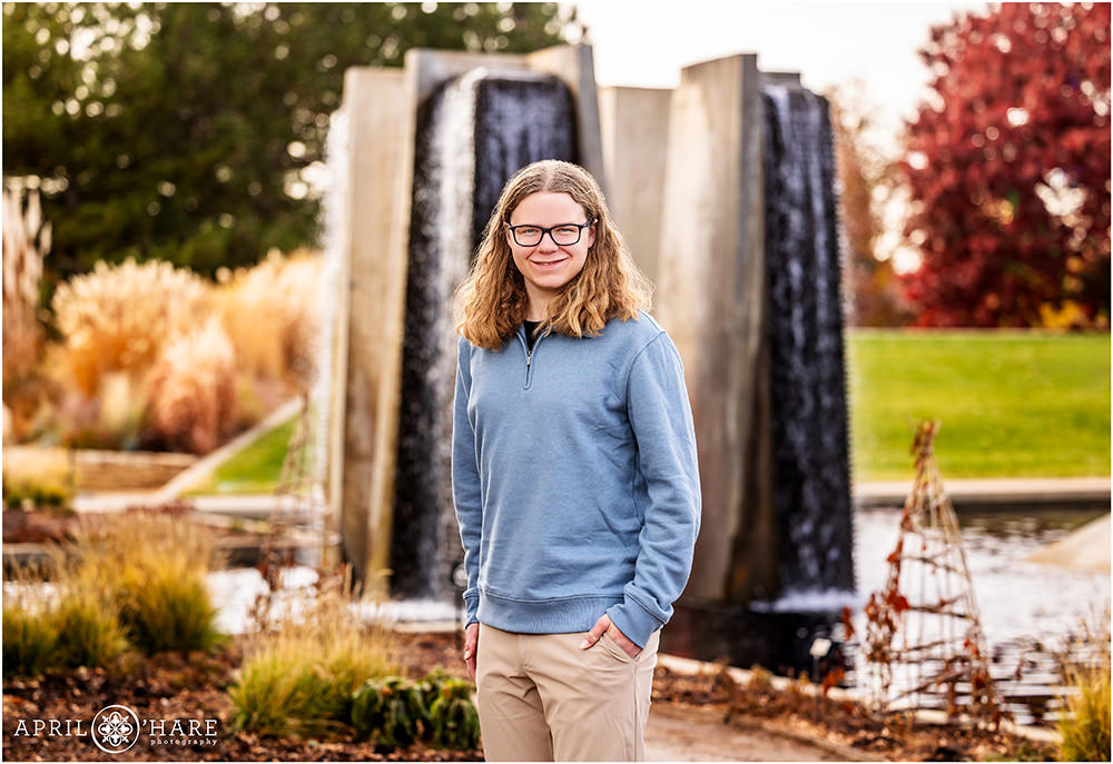 High school senior boy wearing a blue long sleeved shirt stands in front of garden fountain at Denver Botanic Gardens during November