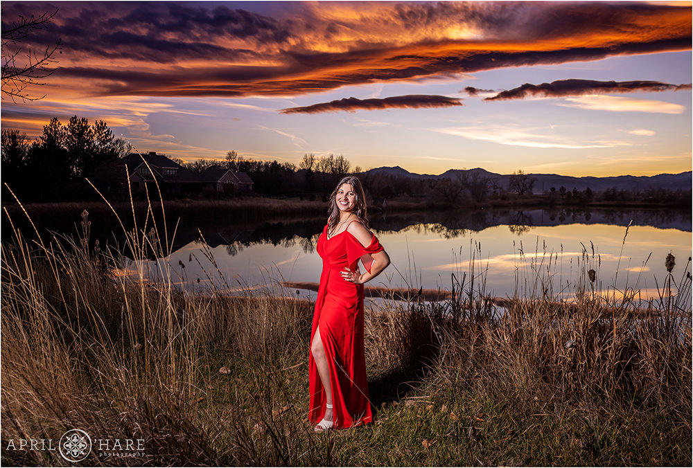 High school senior girl wearing a long red dress poses in front of a sunset mountain lake backdrop at home in Colorado