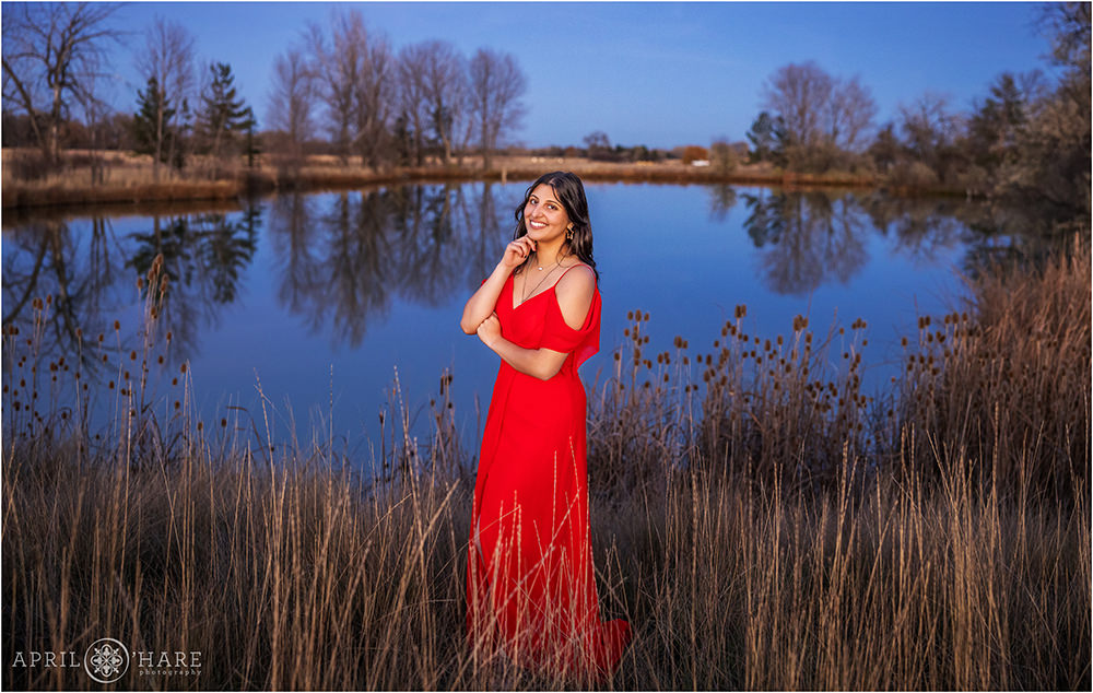 Senior girl wearing a bright red dress poses in front of a small lake next to her home in Colorado