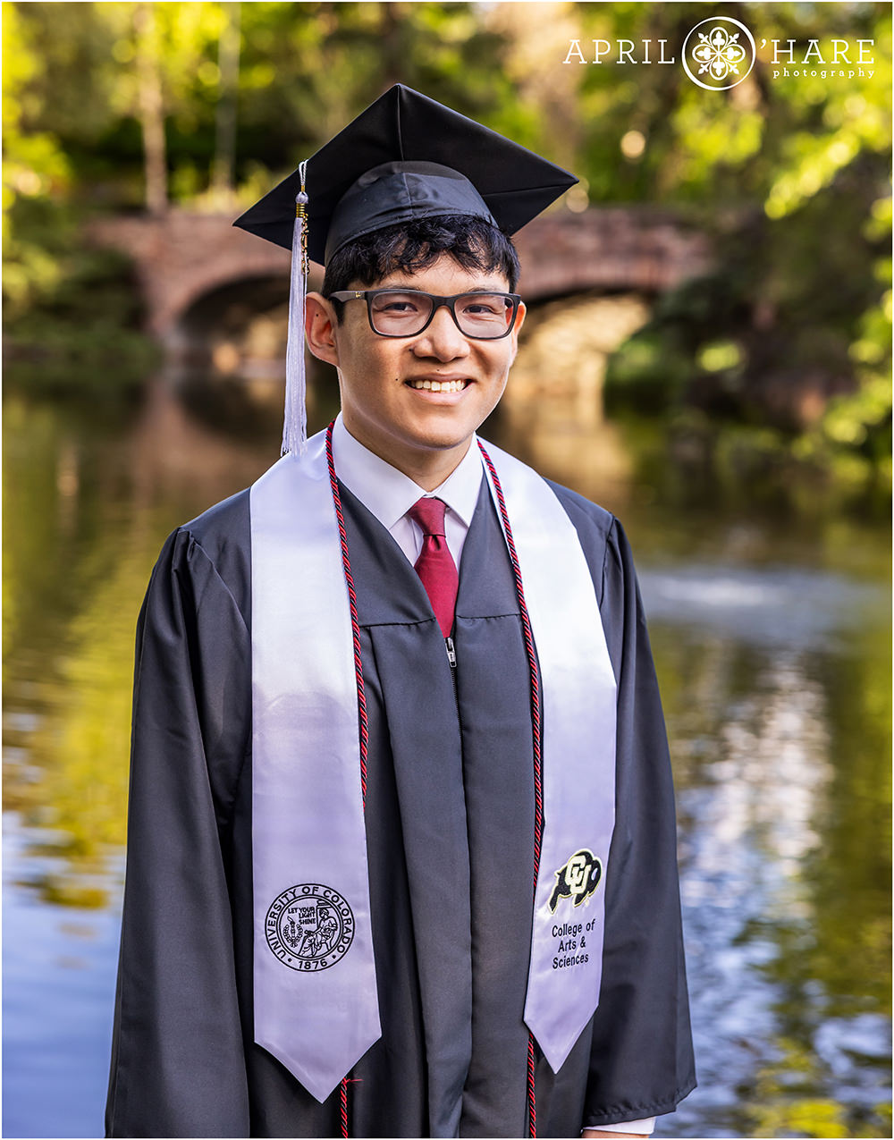 Cap and gown graduation portrait at Varsity Lake with the bridge in the backdrop at University of Colorado during spring